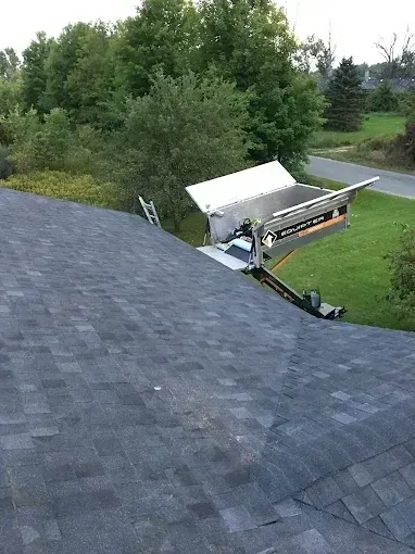 View from a shingled roof onto a backyard with trees, lawn, and a white trailer near a driveway