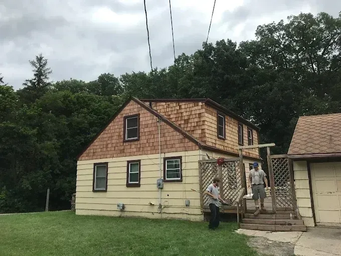 Small beige house with a steep roof, a side porch, and trees behind it