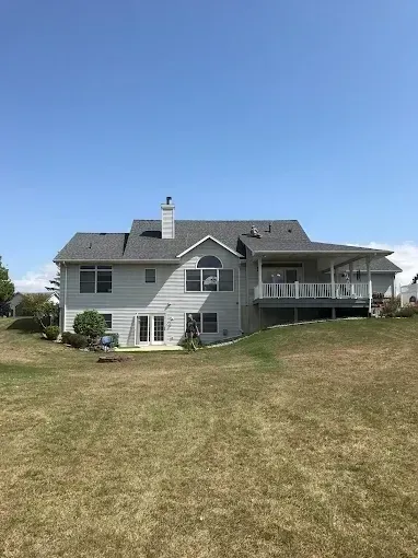 Large gray two-story house with a deck on a grassy hill under a clear blue sky