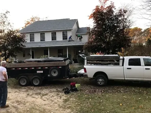 White pickup and dump trailer parked in front of a two-story house with a person standing nearby.