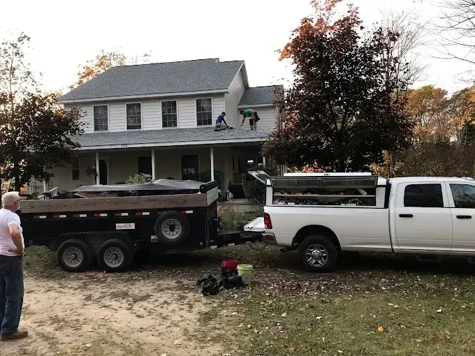 Two trucks and a dump trailer in front of a gray house, with a man standing at left