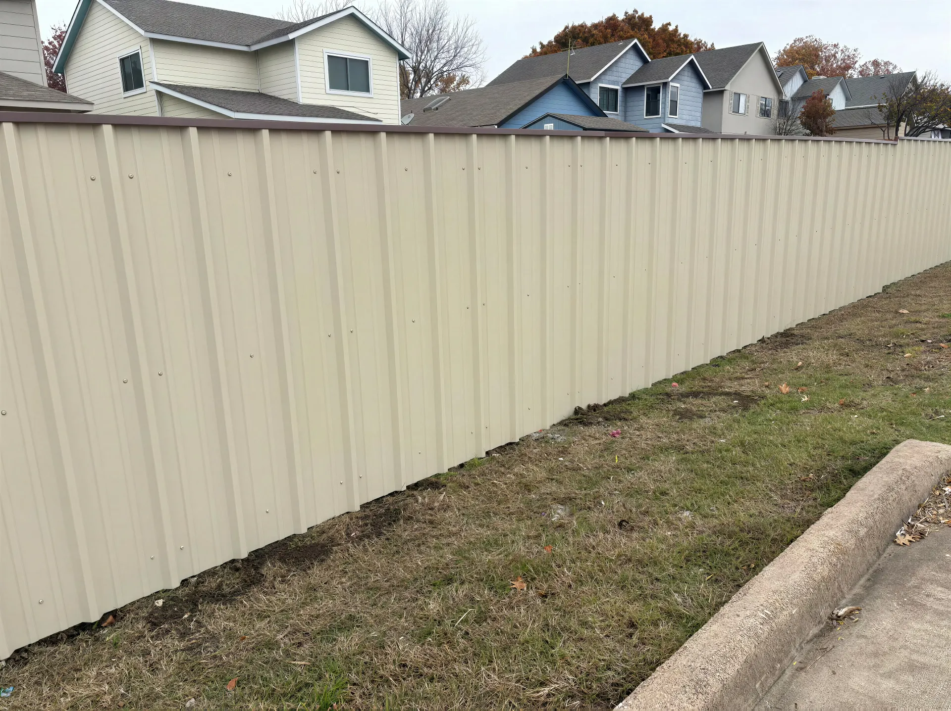 Beige vinyl fence along a grassy roadside with houses visible behind it.