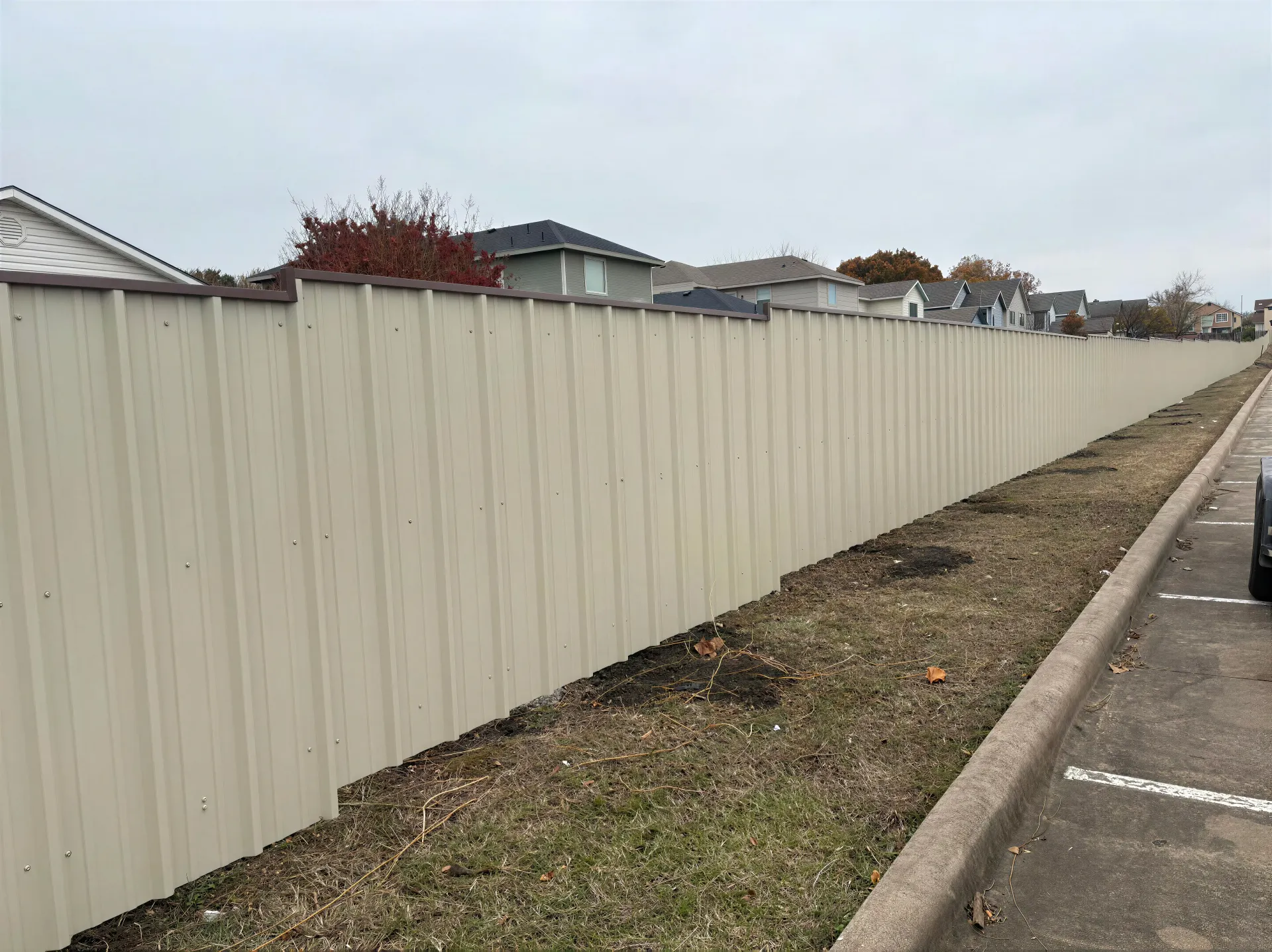 Long beige corrugated fence beside a narrow gravel strip and roadside parking under an overcast sky