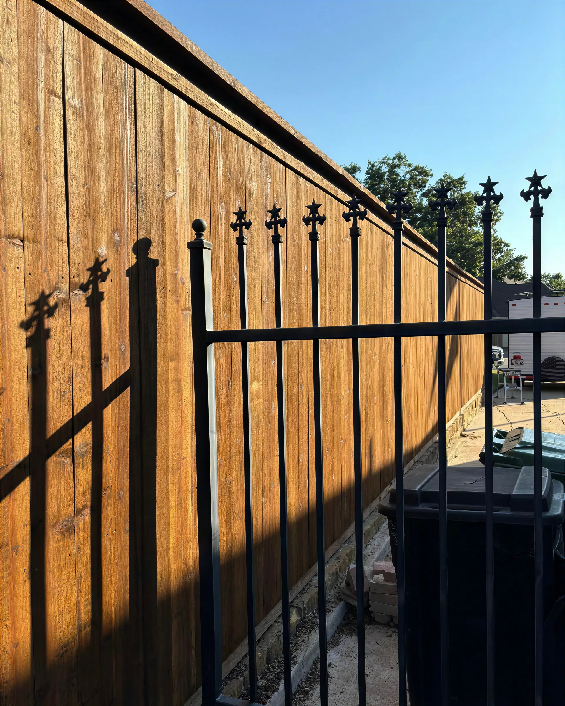 Wooden fence with black metal spikes casting long shadows beside a driveway and trash bin.