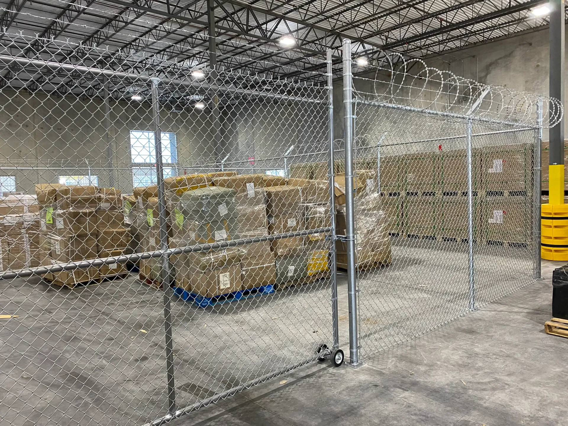 Chain-link gate in a warehouse aisle, with palletized boxes and yellow equipment in the background
