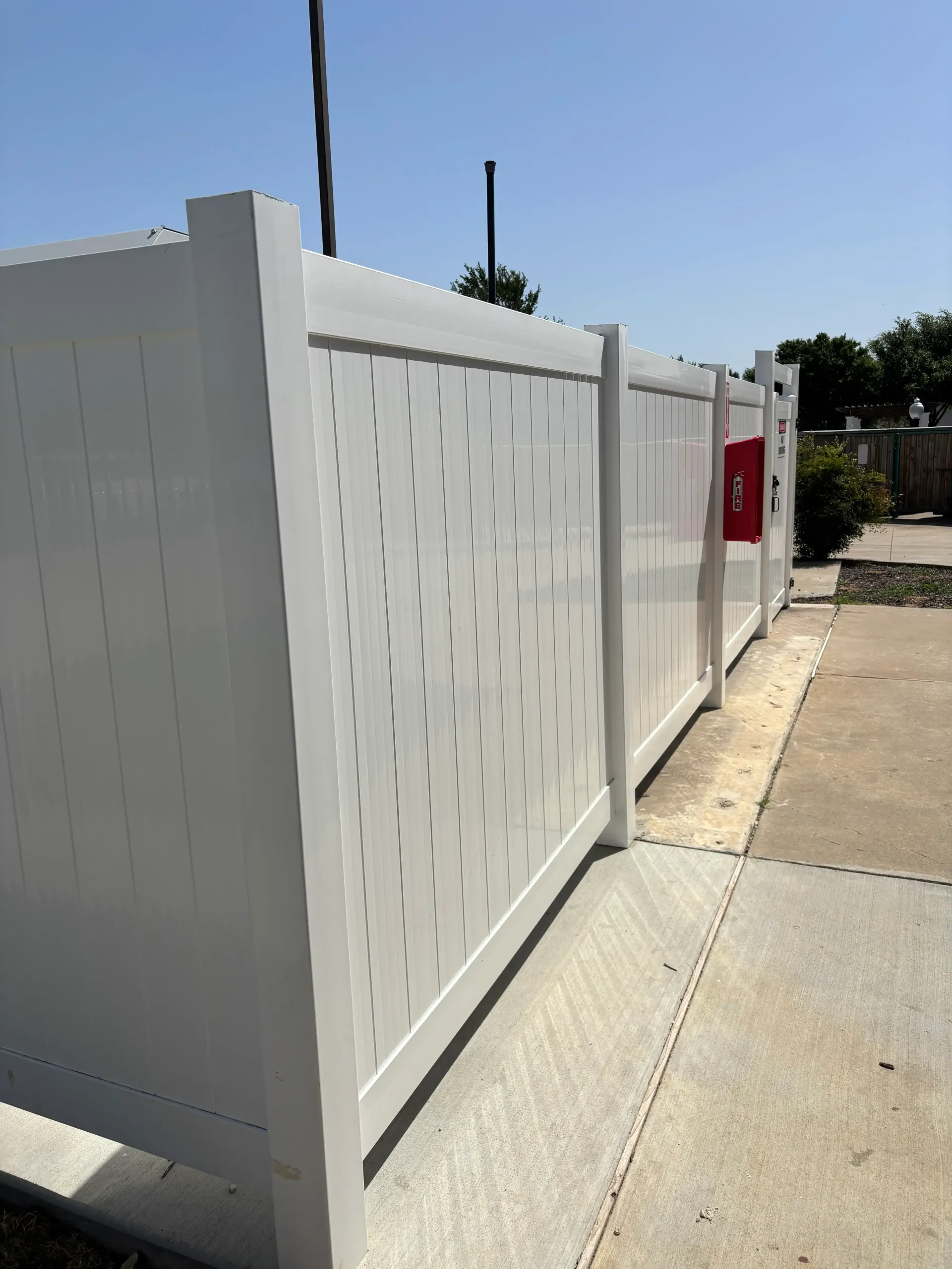 White fence along a sidewalk beside a street, with a red sign on the gate.