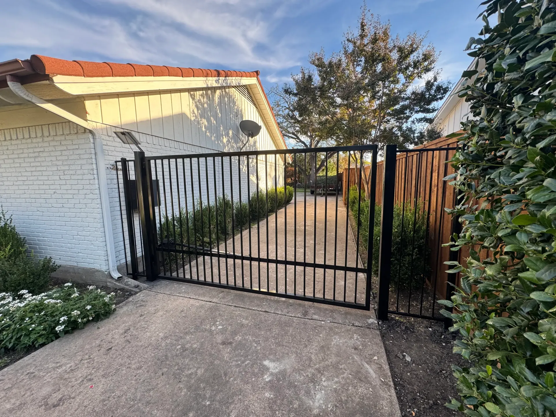 Black metal gate across a driveway beside a white brick house and garden path