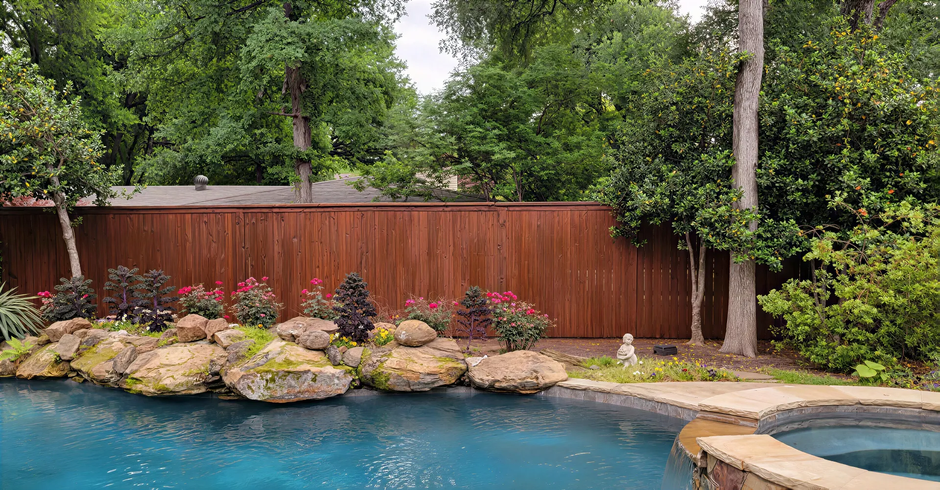 Backyard pool with turquoise water, stone coping, rock border, wooden fence, and lush trees.