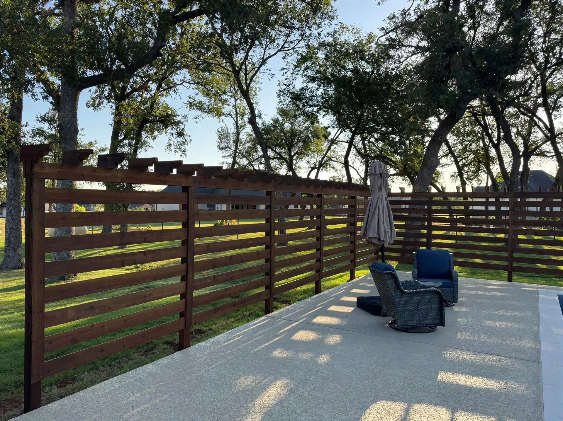 Backyard patio with black chairs, closed umbrella, and wooden fence under trees in sunlight