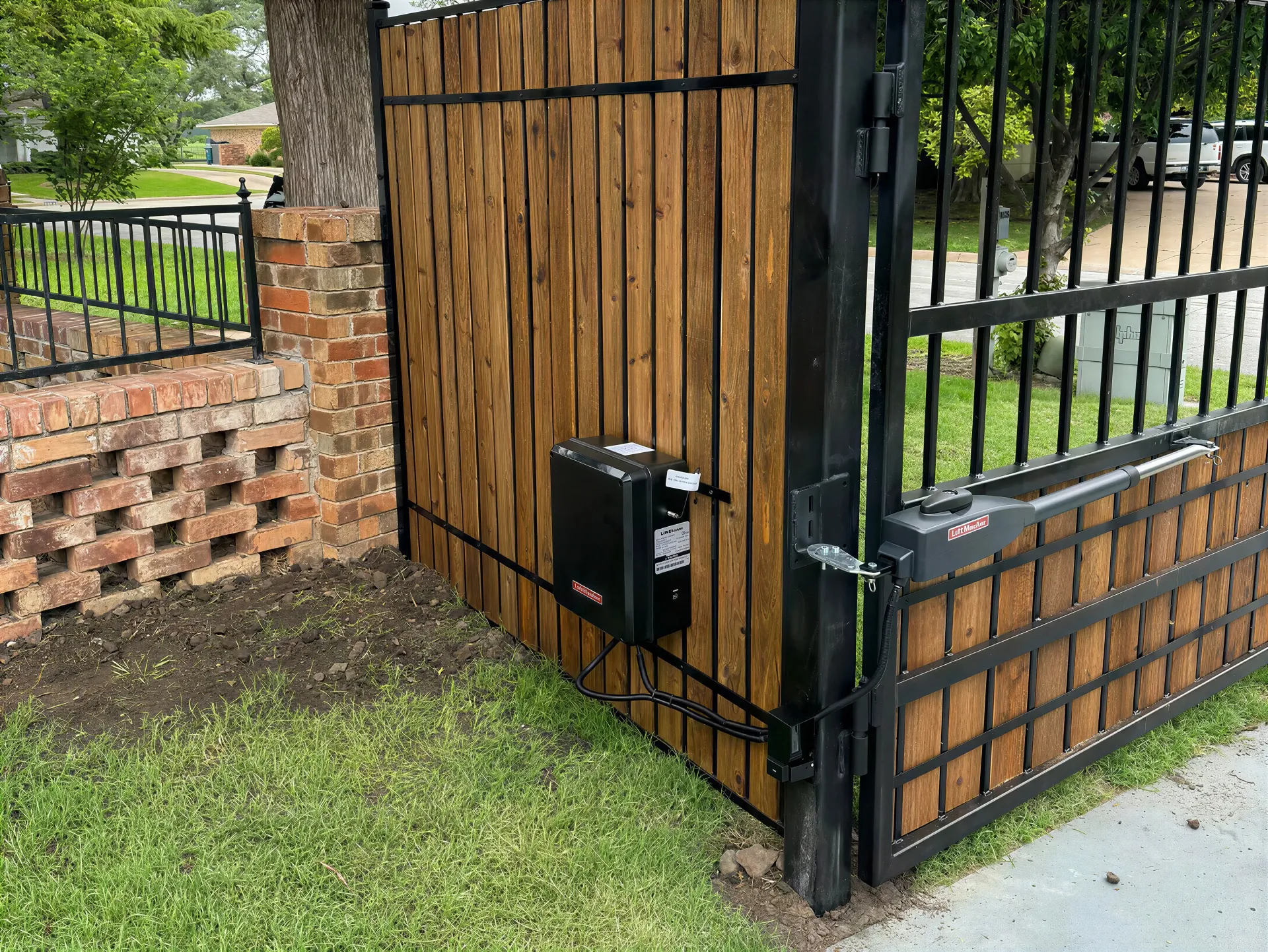 Black metal gate beside a wooden fence and stone wall, with a small black box mounted near the corner.