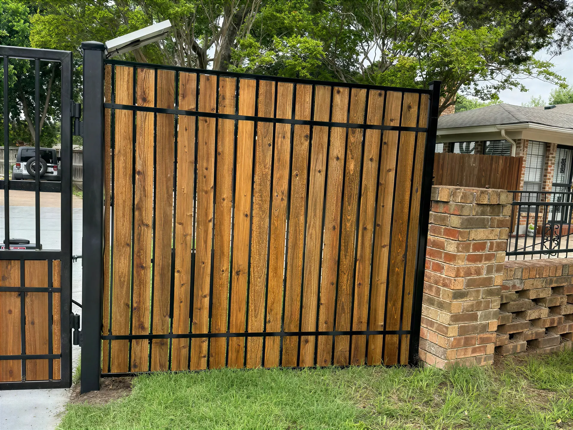 Wooden gate beside a brick wall and black metal fence, with grass and trees in the background.