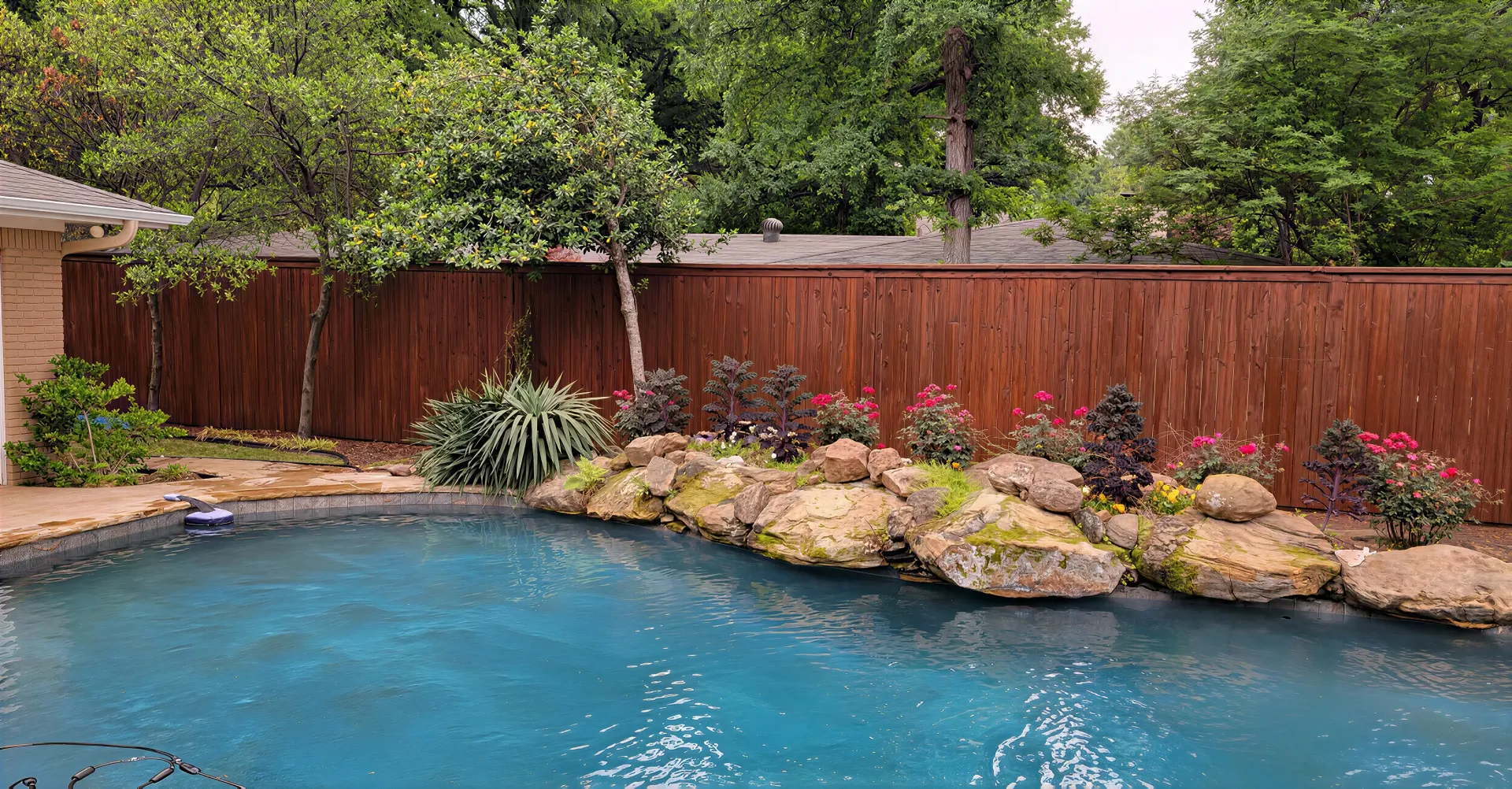 Brown wooden fence beside gravel path under leafy trees in a sunny yard