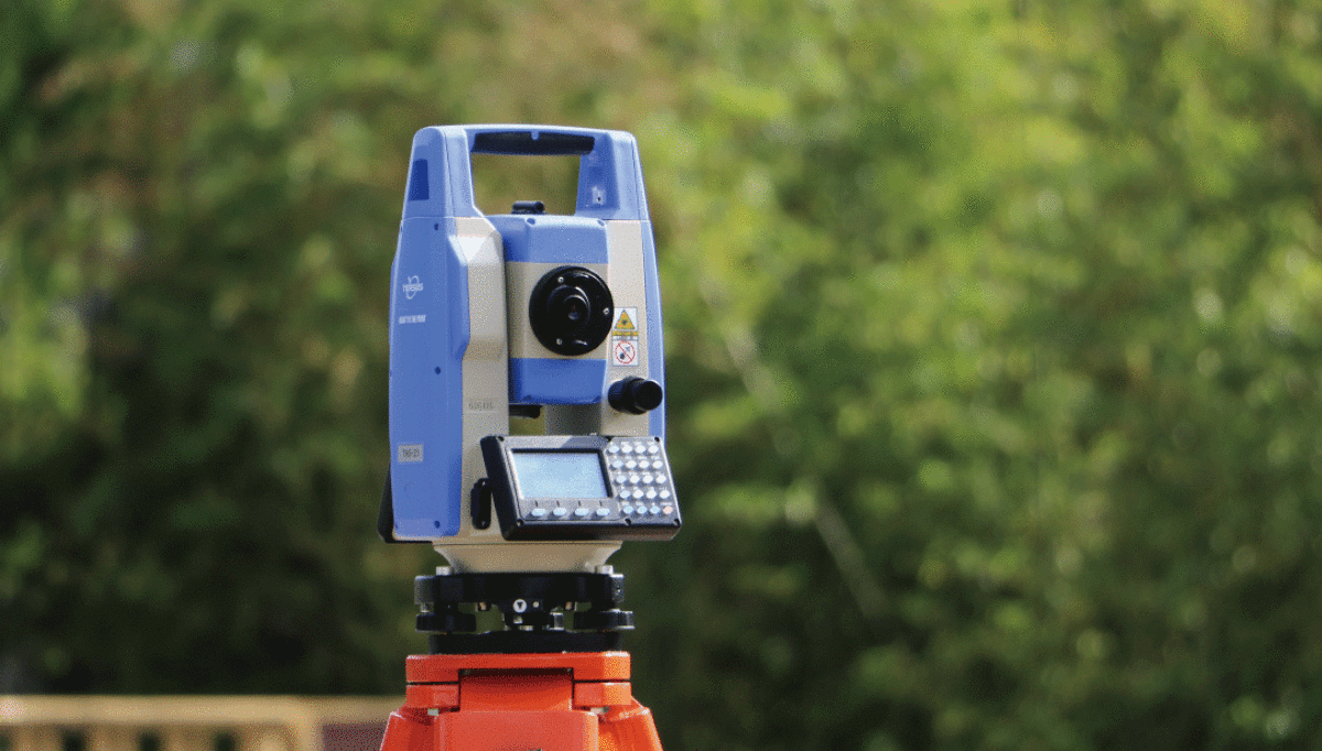 Blue surveying total station on an orange tripod against a blurred green outdoor background