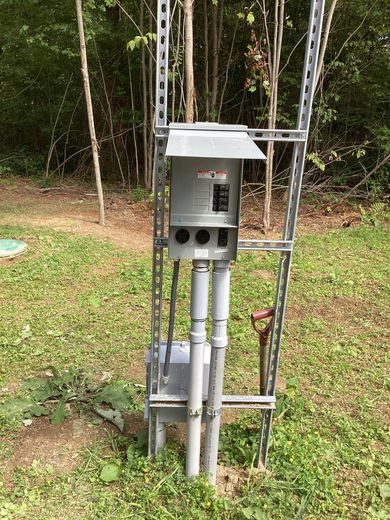 Outdoor electrical meter and breaker box mounted on a metal frame beside a wooded area.