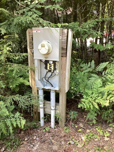 Outdoor electrical meter box mounted on posts in a wooded area, with pipes and foliage around it