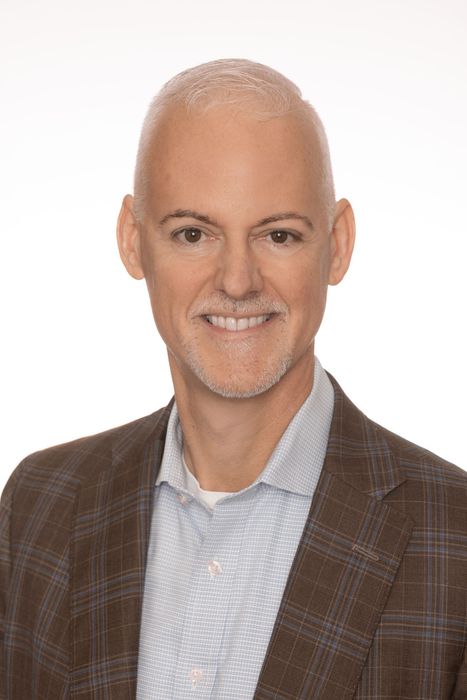 Studio portrait of a smiling bald man in a brown plaid blazer over a light blue shirt on a white background