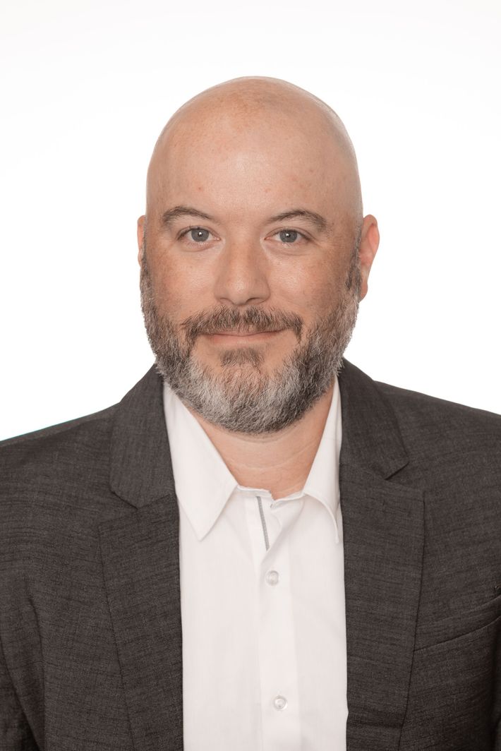 Studio headshot of a bald man with a gray beard wearing a dark blazer and white shirt on a white background