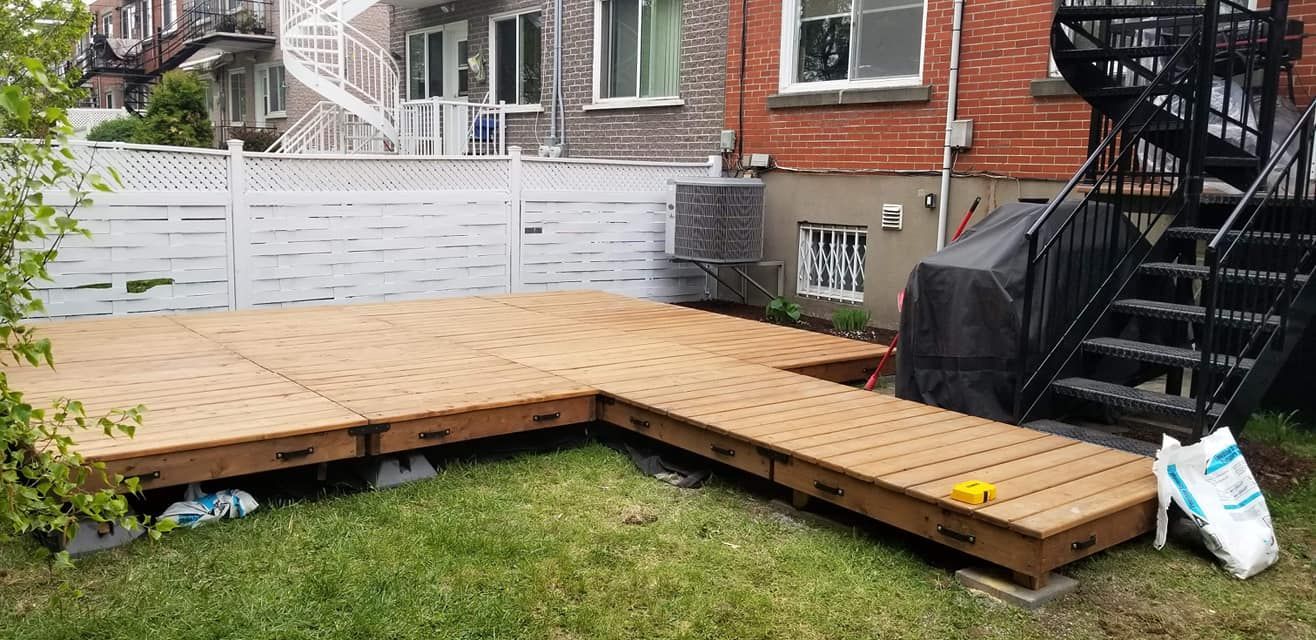 Terrasse en bois dans un jardin avec une clôture blanche, un bâtiment en briques rouges et un escalier en colimaçon.