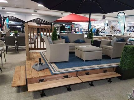 Mobilier de jardin avec canapé, chaises et parasol sur une terrasse en bois. Des bancs sont disposés devant.