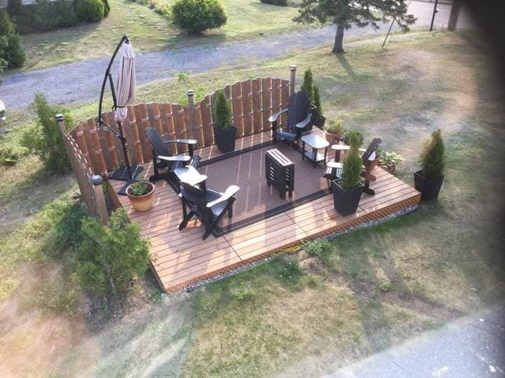 Terrasse en bois avec chaises Adirondack, petite table, parasol, entourée d'une clôture et de verdure.