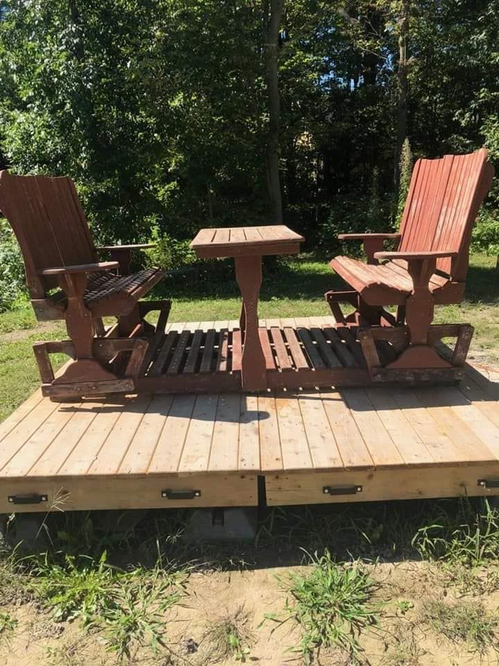 Fauteuil à bascule double en bois avec une petite table, peint en rouge, sur une plateforme en bois à l'extérieur.