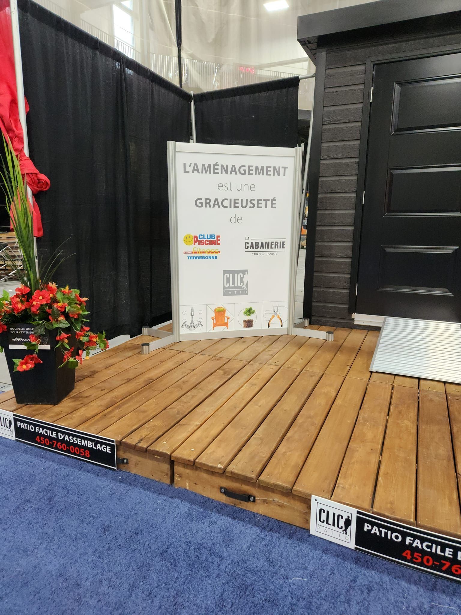 Présentoir de terrasse en bois lors d'un salon professionnel, avec une pancarte indiquant « L'Aménagement de la Rusetre ». Porte noire et fleurs.