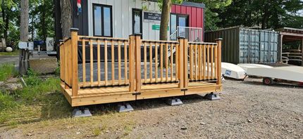 Terrasse en bois avec rambarde sur gravier, devant un bâtiment blanc aux finitions noires.