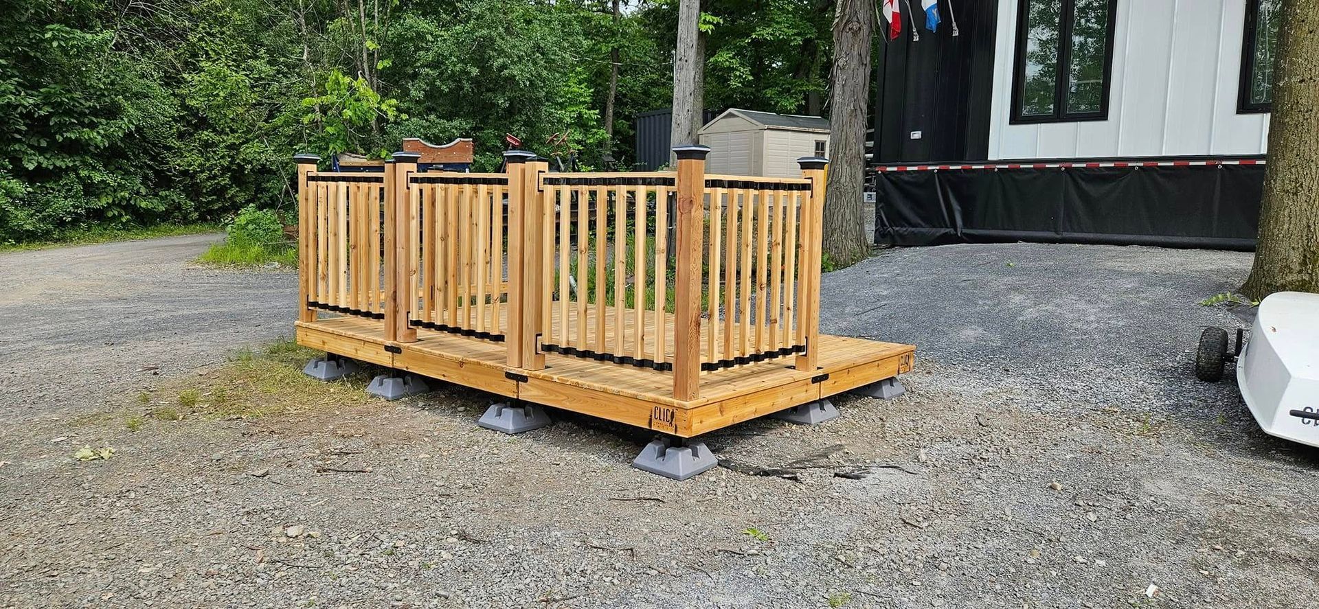 Terrasse en bois avec rambarde sur gravier, à côté d'un bâtiment et d'arbres.