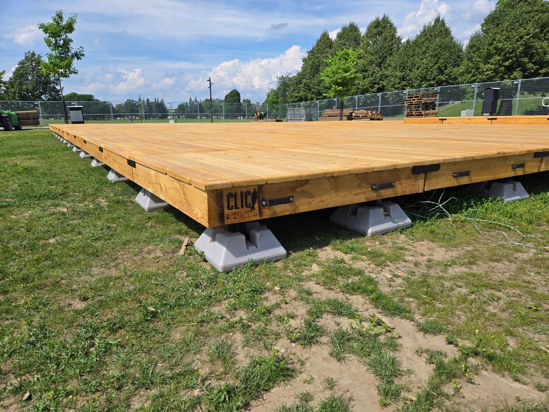 Plateforme en bois sur blocs de béton dans un champ herbeux, probablement pour un chantier. Cadre extérieur ensoleillé.