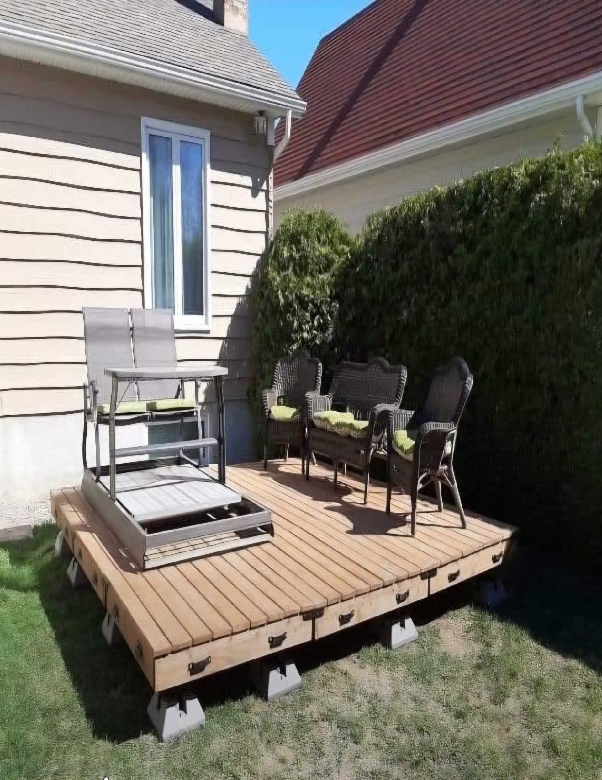 Terrasse en bois avec mobilier de jardin sur une pelouse, à côté d'un bâtiment et d'une haie.