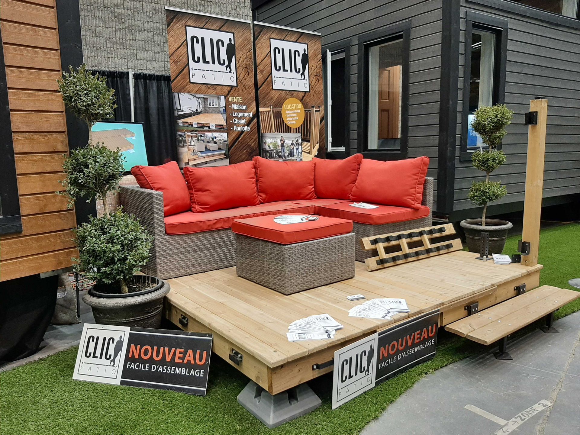 Aménagement d'un coin salon extérieur avec coussins rouges, petits arbres, terrasse en bois et panneau « CLIC ».