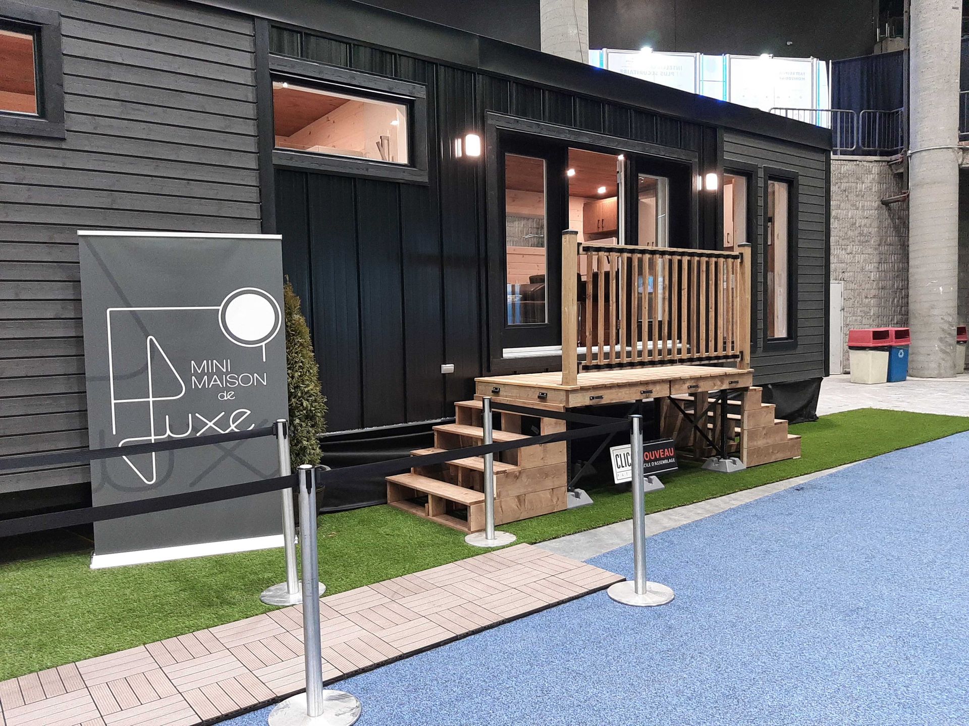 Petite maison noire avec terrasse en bois et enseigne « Luxe ». Sur gazon artificiel à sol bleu.