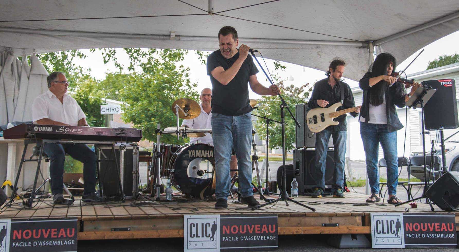 Groupe de musique jouant sur scène sous un chapiteau : chanteur, claviériste, batteur, bassiste, violoniste. Cadre extérieur.
