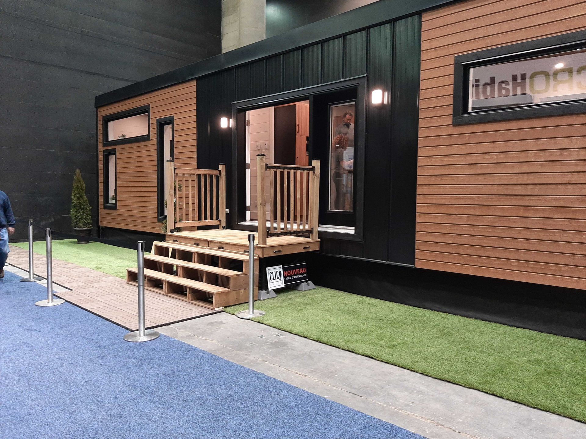 Petite maison avec façade en bois, rampe d'accès. Bardage noir, deux fenêtres, pelouse et moquette bleue.