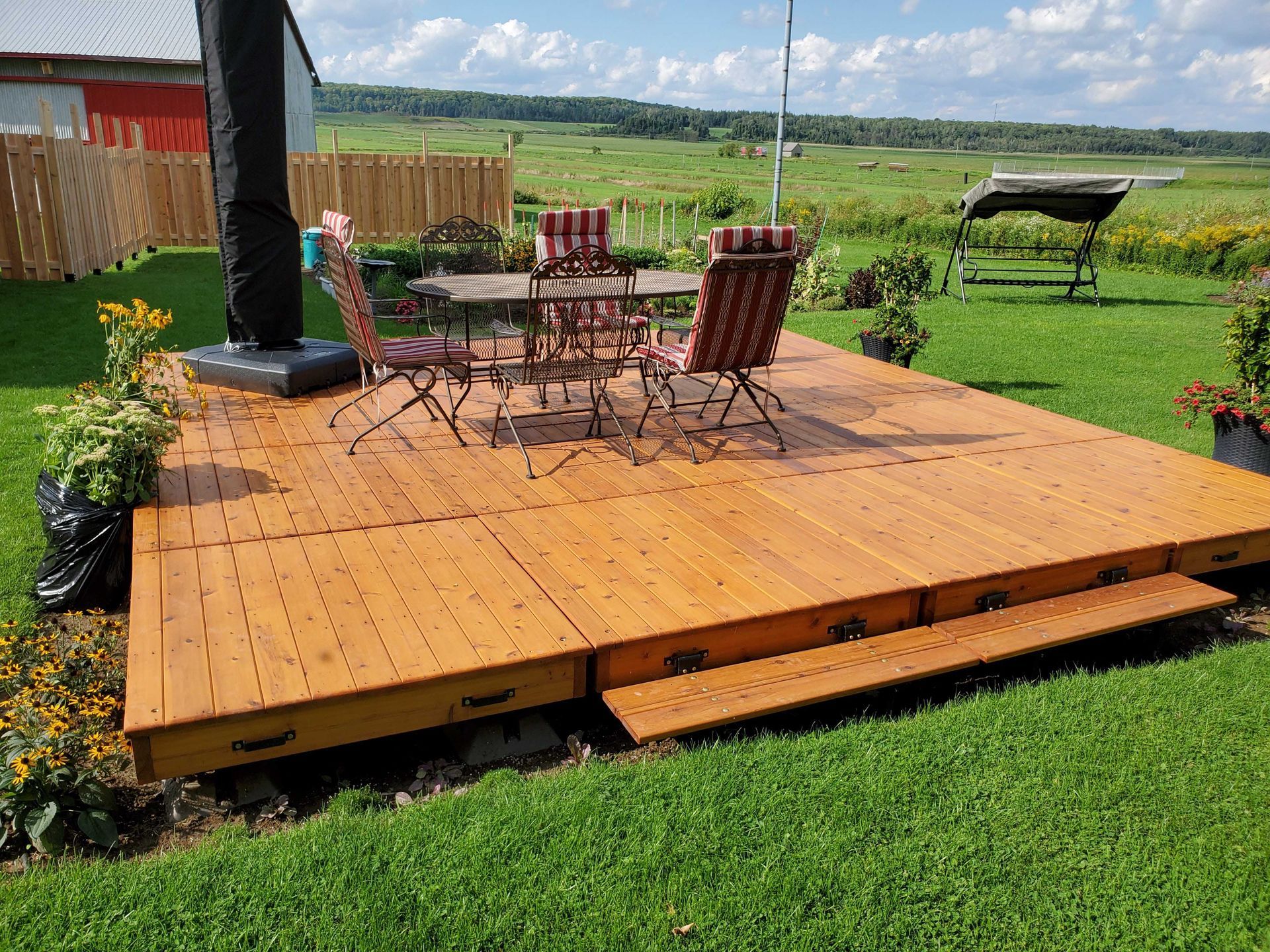 Terrasse en bois avec table et chaises sur une pelouse ; un champ se dessine à l'arrière-plan.