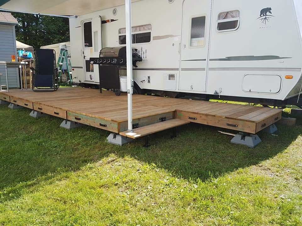 Terrasse en bois avec marches à côté d'un camping-car blanc, sur un emplacement de camping herbeux avec un barbecue.