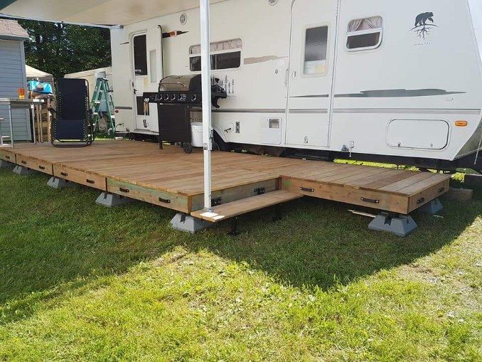 Terrasse en bois avec marches jouxtant un camping-car blanc, sur une pelouse. Un barbecue noir est visible.