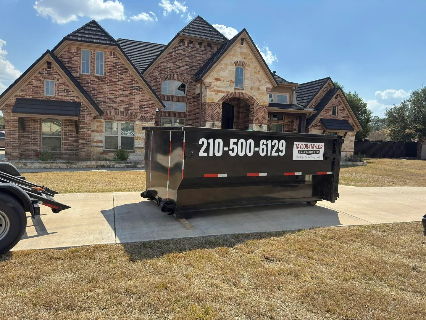 A large black dumpster is parked in front of a brick house.