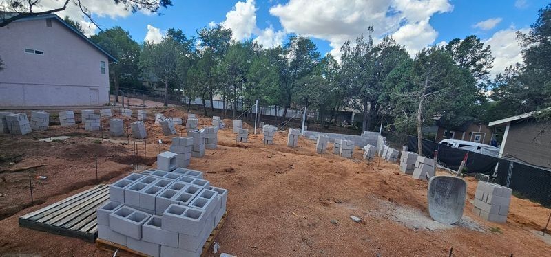 Construction site with stacked cinder blocks and concrete pillars on a cleared dirt lot under a blue sky