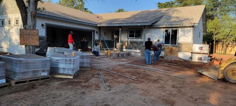 Construction workers on a muddy driveway outside a house with stacked pavers and building materials.