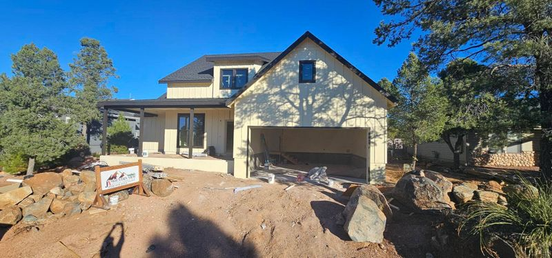 New beige house under construction with a garage, dirt foreground, and trees under a clear blue sky