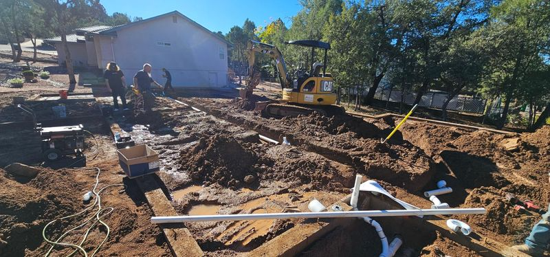 Construction site with excavated dirt, workers, and a yellow truck near a house under clear skies