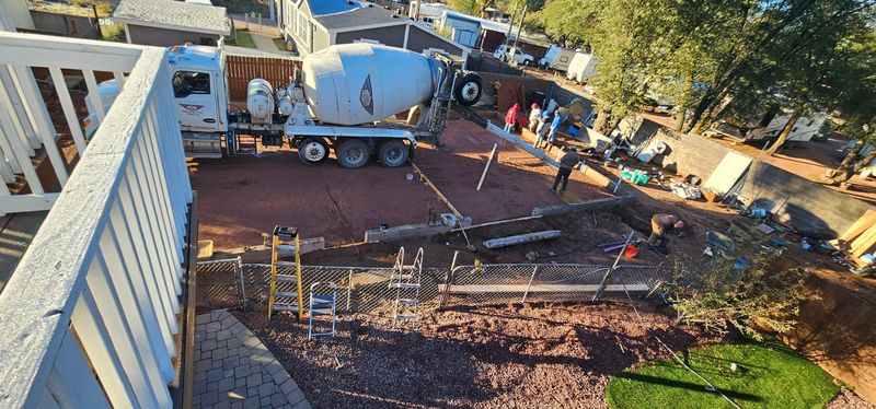 Construction crew pouring a concrete driveway beside a white house, with a cement truck and workers in a sunny yard