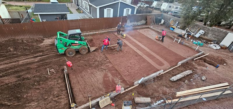 Aerial view of a backyard excavation site with a green digger and workers leveling reddish soil.