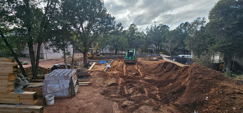 Construction site with excavator digging a large dirt mound near trees and stacked materials