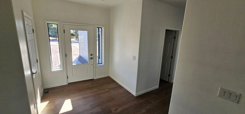 Empty room with dark wood floors, white walls, and front glass doors letting in sunlight.