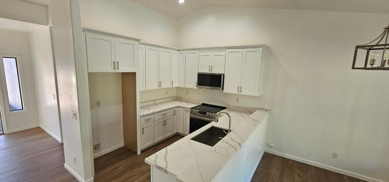 Bright white empty kitchen with island, black appliances, and wood flooring, viewed from the living area