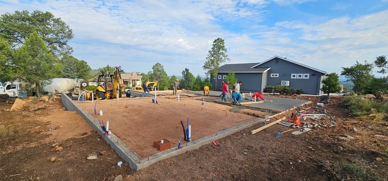 Construction site with workers pouring a concrete foundation beside a blue house and trees