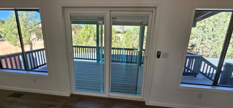Interior view of a sliding glass door opening to a wooden deck and outdoor trees, flanked by two windows.