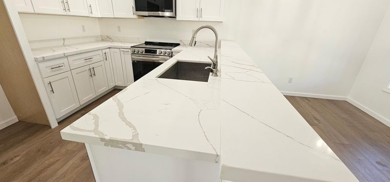 Bright white kitchen with marble island, white cabinets, stainless appliances, and a sink near a window